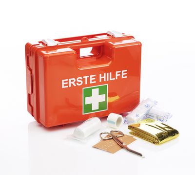 Kleiner Koffer in orange mit der Aufschrift "Erste Hilfe".  Vor dem Koffer liegen eine Schere, Pflaster, Binden und eine Rettungsdecke