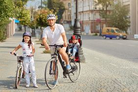Ein Mädchen im Grundschulalter hat einen Fahrradhelm auf und steht neben einem Fahrrad. Ein Mann, ebenfalls mit Helm steht mit seinem Rad auf einem Fußweg. Er hat ein Kleinkind mit Helm im Kindersitz.