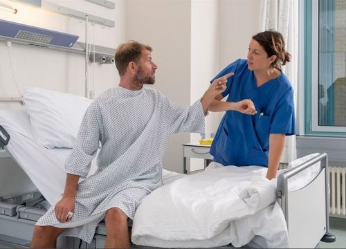 Ein männlicher Patient sitzt auf einem Krankenhausbett und beschimpft eine neben dem Bett stehende Krankenschwester