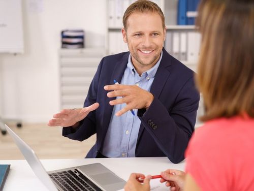 An einem Tisch sitzen ein Mann und eine Frau und auf dem Schreibtisch liegt ein Laptop. Der Mann sagt mit einem freundlichen Gesicht etwas zu der Frauerzählt mit