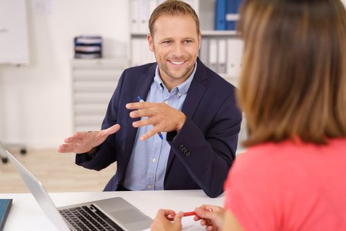 An einem Tisch sitzen ein Mann und eine Frau und auf dem Schreibtisch liegt ein Laptop. Der Mann sagt mit einem freundlichen Gesicht etwas zu der Frauerzählt mit