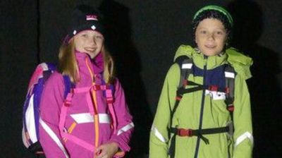 Ein Mädchen mit einer rosa Jacke und ein Junge mit einer grünen Jacke im Dunkeln. Die Jacken reflektieren.