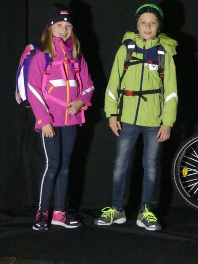 Ein Mädchen mit einer rosa Jacke und ein Junge mit einer grünen Jacke im Dunkeln. Die Jacken reflektieren.