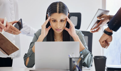 Fotografie einer gestressten Frau am Schreibtisch mit Laptop, umgeben von mehreren Personen, die Tablets und Smartphones halten. Darstellung von Arbeitsdruck und Multitasking in einem modernen Büro.