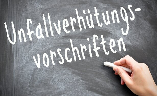 Auf einer Tarif wird von einer Menschenhand mit weißer Kreide das Wort Unfallverhütungsvorschriften geschrieben