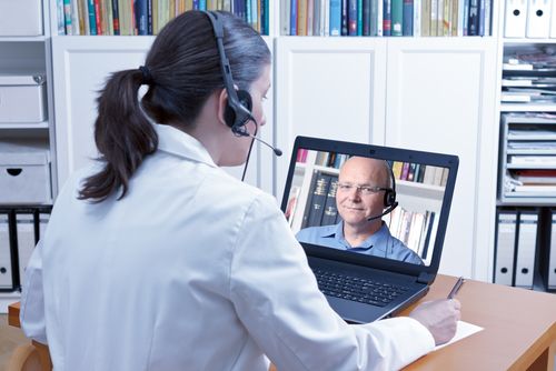 Eine Ärztin sitzt mit Kopfhörer vor einem Laptop, auf dem Laptop ist ein Mann zu sehen, der auch einen Kopfhörer trägt