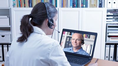 Eine Ärztin sitzt mit Kopfhörer vor einem Laptop, auf dem Laptop ist ein Mann zu sehen, der auch einen Kopfhörer trägt