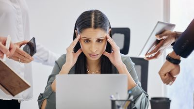 Eine Frau sitzt vor einem Laptop, stützt ihren Kopf mit den Händen. Links und rechts von der Frau halten ihr fremde Hände Handys, Laptops und Uhren entgegen