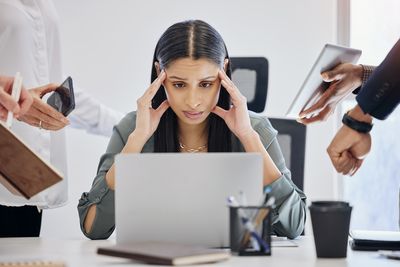Eine Frau sitzt vor einem Laptop, stützt ihren Kopf mit den Händen. Links und rechts von der Frau halten ihr fremde Hände Handys, Laptops und Uhren entgegen