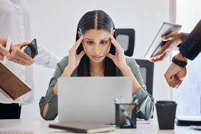 Eine Frau sitzt vor einem Laptop, stützt ihren Kopf mit den Händen. Links und rechts von der Frau halten ihr fremde Hände Handys, Laptops und Uhren entgegen
