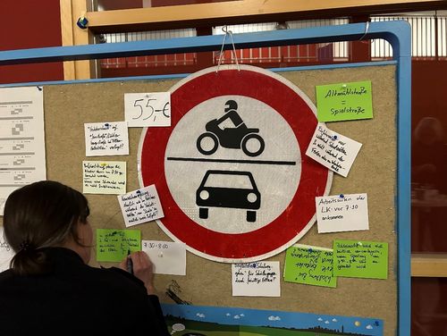 Eine Frau hängt einen Zettel an eine Pinnwand, an der auch ein Verkehrsschild hängt.