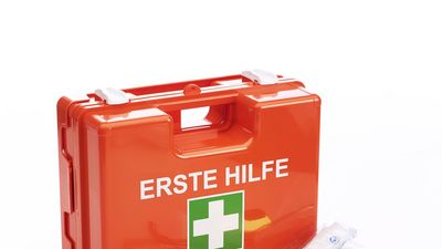 Kleiner Koffer in orange mit der Aufschrift "Erste Hilfe".  Vor dem Koffer liegen eine Schere, Pflaster, Binden und eine Rettungsdecke