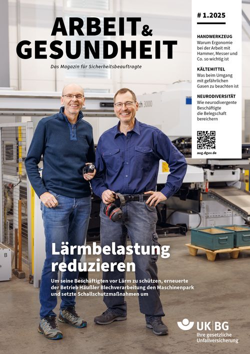 Zwei Männer stehen nebeneinander in einer Werkstatt und schauen lachend in eine Kamera.