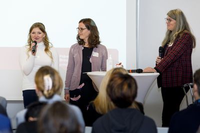 Eine Schülerin hält über Mikrofon einen Vortrag, zwei Frauen stehen am Stehtisch neben ihr in einem Saal