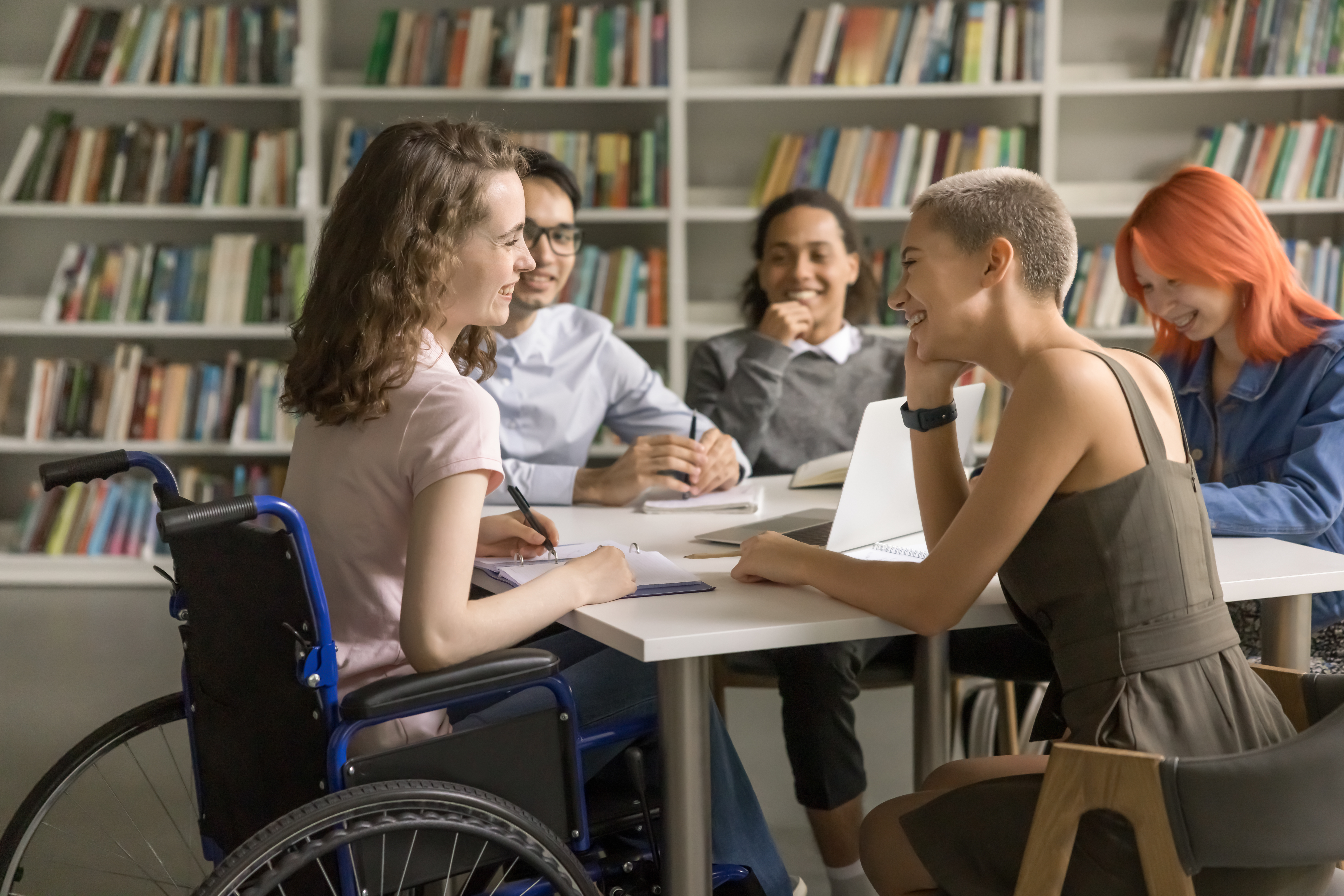Fünf Jugendliche, davon sitzt ein Mädchen im Rollstuhl, sitzen lachend an einem Tisch