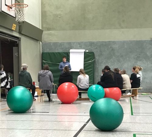 In einer Turnhalle sitzen mehrere Menschen auf Stühlen, Bänken und Bällen im Kreis