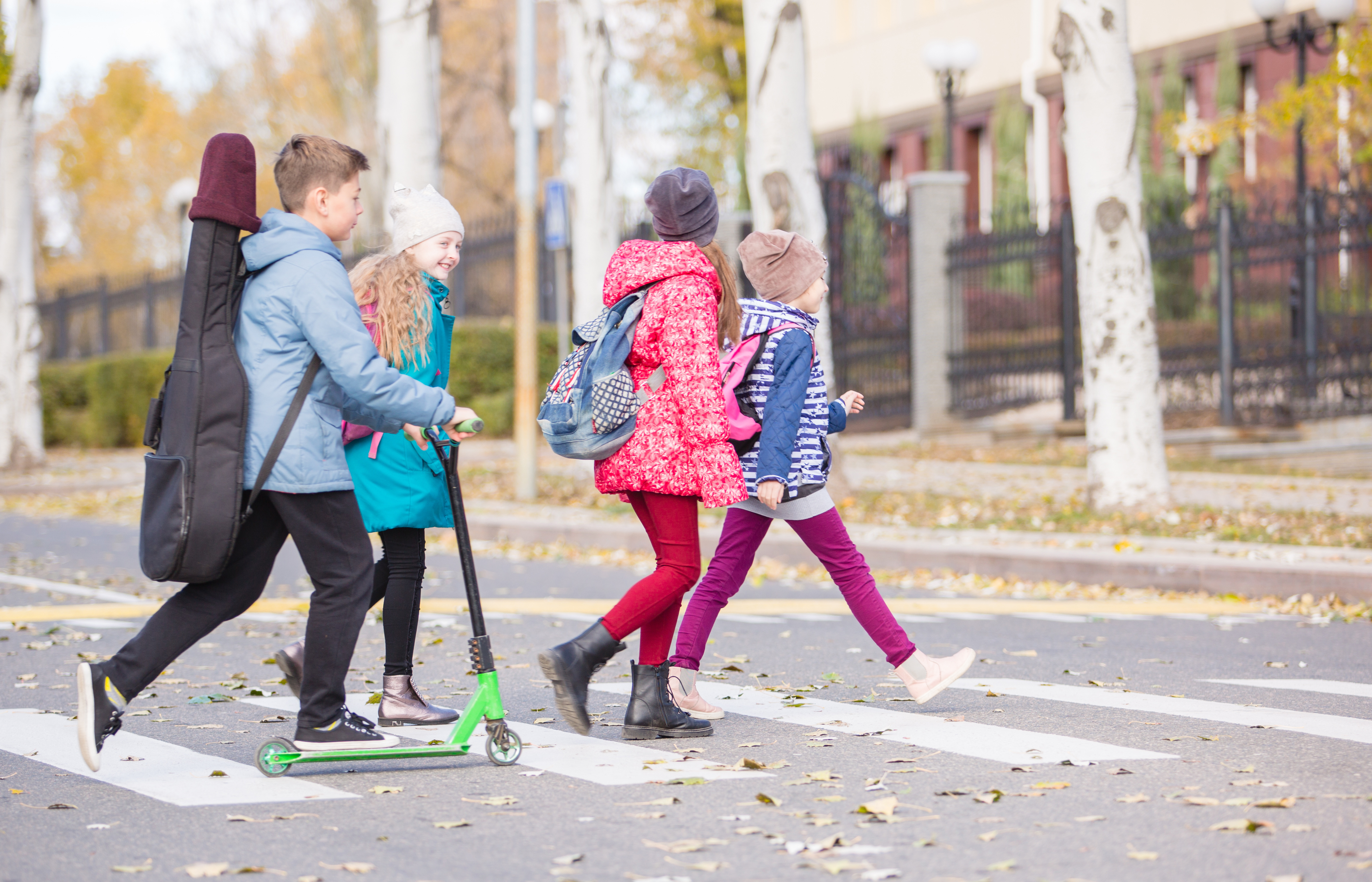 Vier Kinder gehen im Herbst über einen Zebrastreifen. Ein Junge fährt mit einem E-Roller und trägt eine Gitarre auf den Rücken